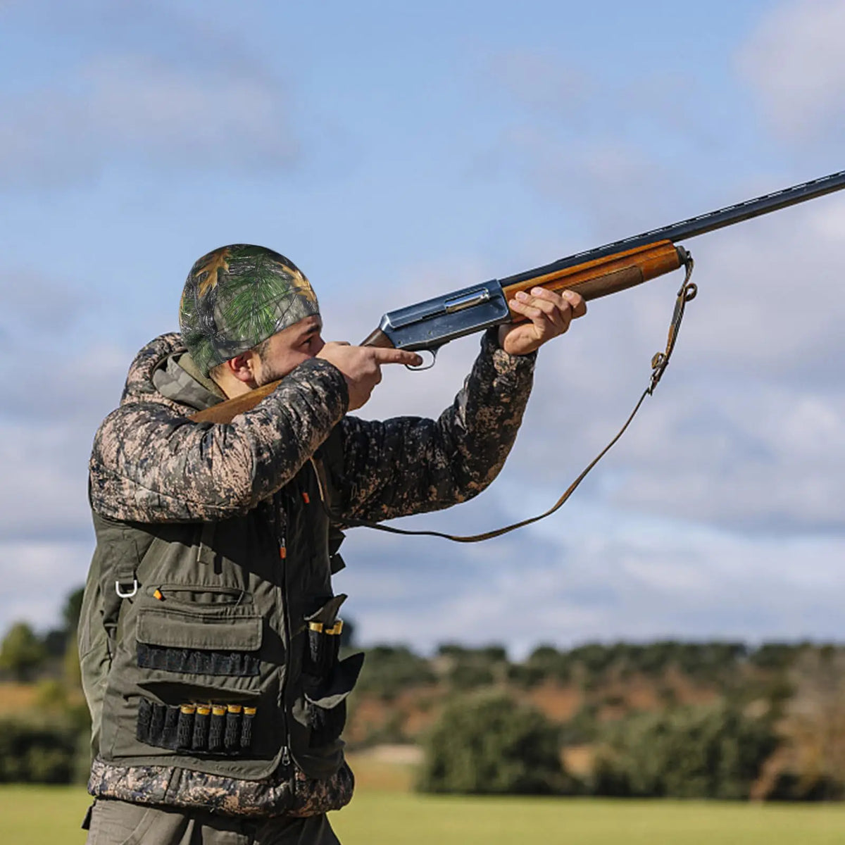 A person wearing a camouflage thermal beanie is aiming a rifle outdoors.