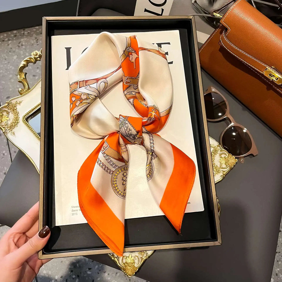 A luxury silk scarf in orange and cream tones is displayed in a black frame next to a gold-edged mirror.