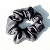 A black silk hair scrunchie with a gathered, ruched texture is shown against a white background.