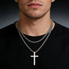 A close-up of a stainless steel cross pendant necklace worn by a man against a black shirt.