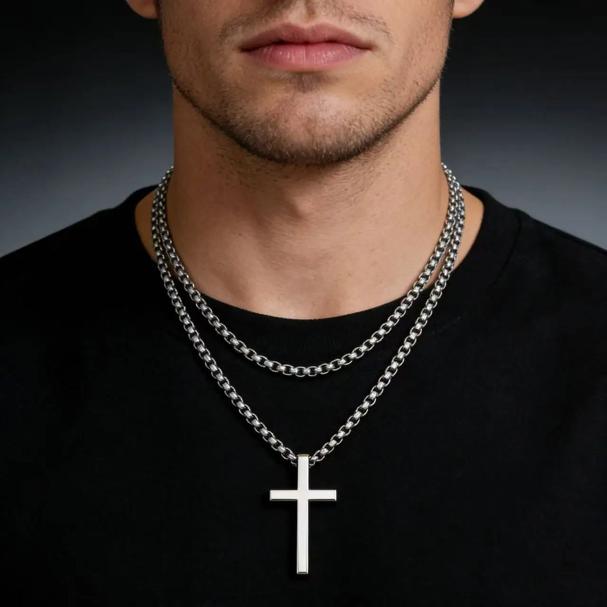 A close-up of a stainless steel cross pendant necklace worn by a man against a black shirt.