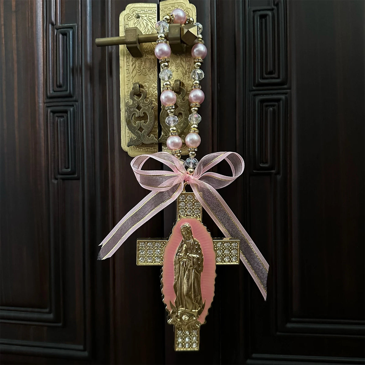 A gold-toned Catholic charm featuring a pink oval with a figure, suspended by a beaded chain and pink ribbon.