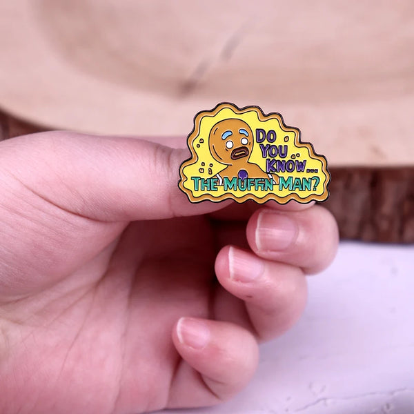 A hand holds an enamel pin featuring a cute cartoon gingerbread man with the text Do you know the muffin man
