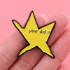A yellow star shaped enamel pin with the text your did it is held in a hand.