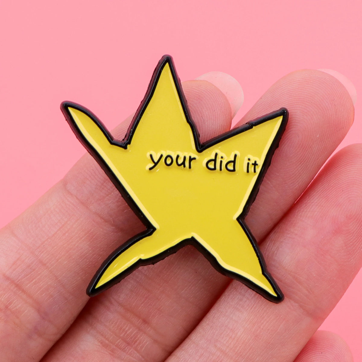 A yellow star shaped enamel pin with the text your did it is held in a hand.
