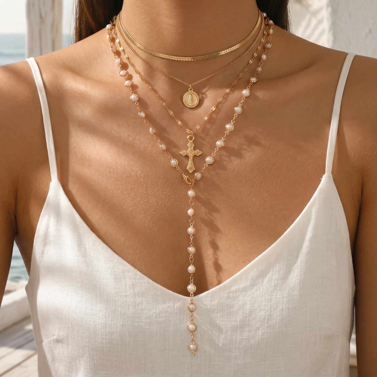 A close-up of a woman wearing a layered gold and pearl cross necklace on her chest.