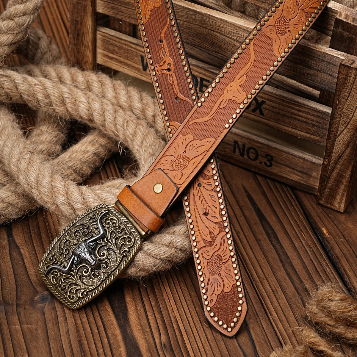 A brown PU leather floral engraved belt with a decorative buckle rests on a wooden surface next to thick rope.