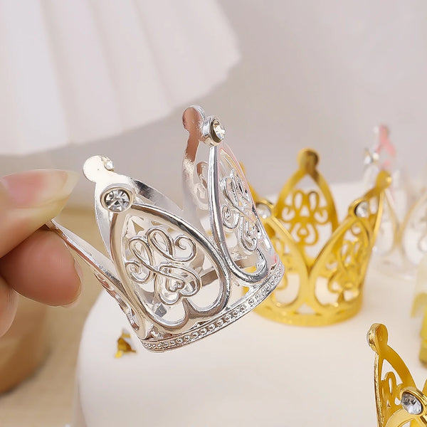 A hand holds a silver and gold mini crown cake topper against a white background.