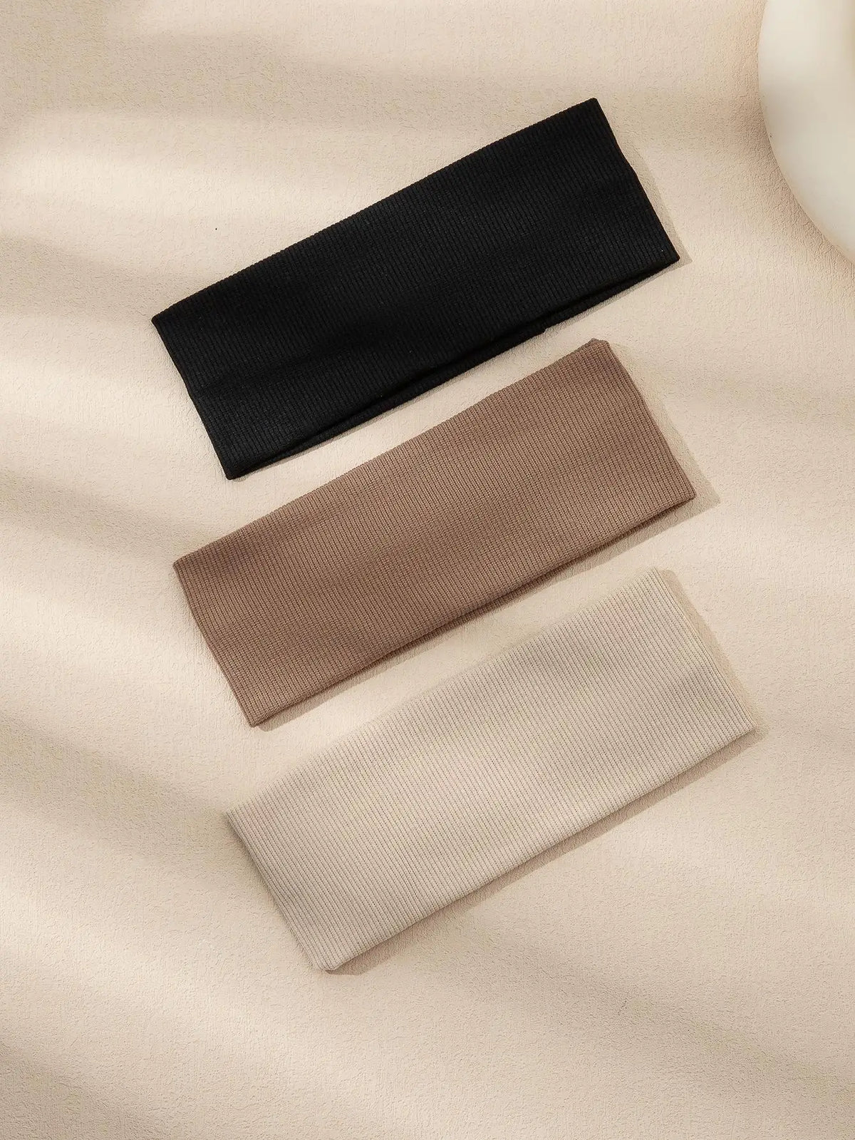 A pack of three solid color fashion headbands in black, brown, and beige laid diagonally on a light fabric background.