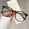 A pair of dark brown, large square anti-blue light spectacles being held against a white background.
