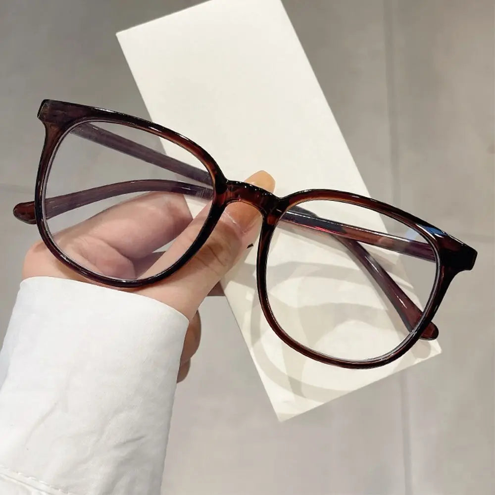 A pair of dark brown, large square anti-blue light spectacles being held against a white background.