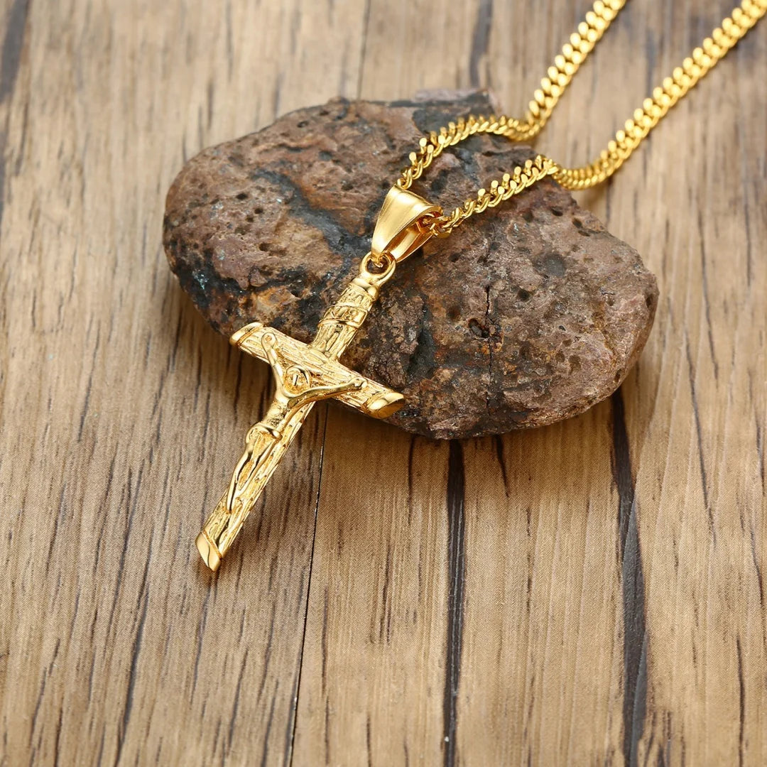 A gold-finished stainless steel cross necklace rests on a dark brown stone against a wooden background.
