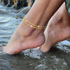 A close-up of a person's foot submerged in water wearing delicate, layered gold glitter anklets.