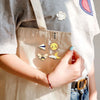 A hand holds a beige canvas tote bag featuring enamel badge pins, including a rose and a sun motif.