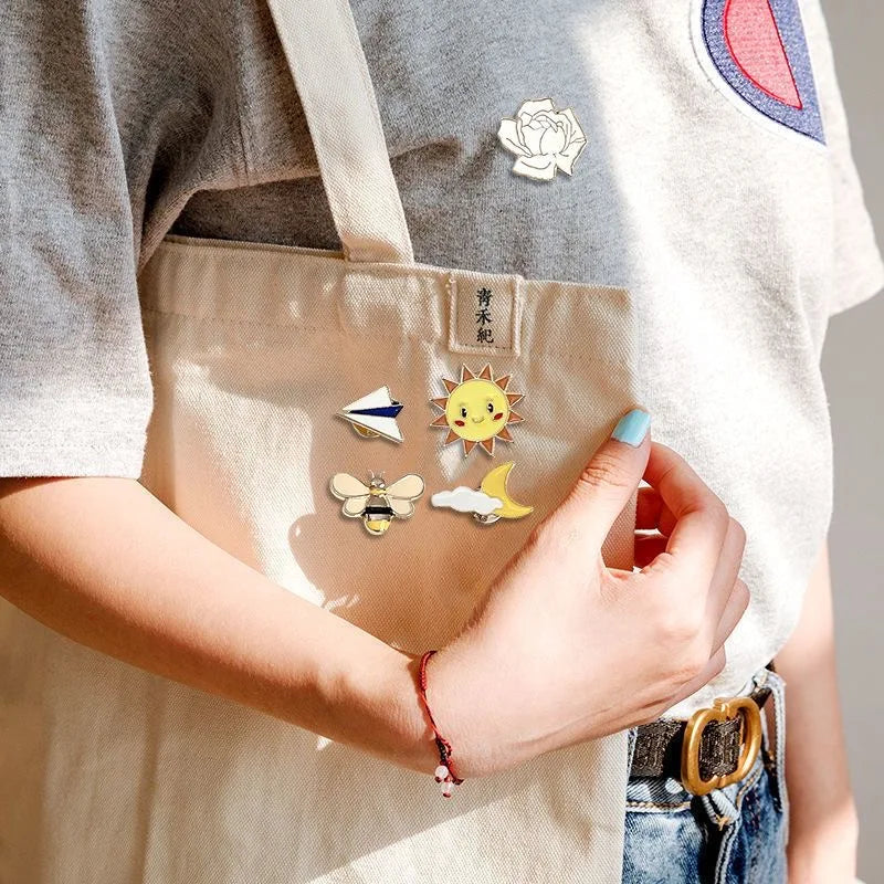 A hand holds a beige canvas tote bag featuring enamel badge pins, including a rose and a sun motif.