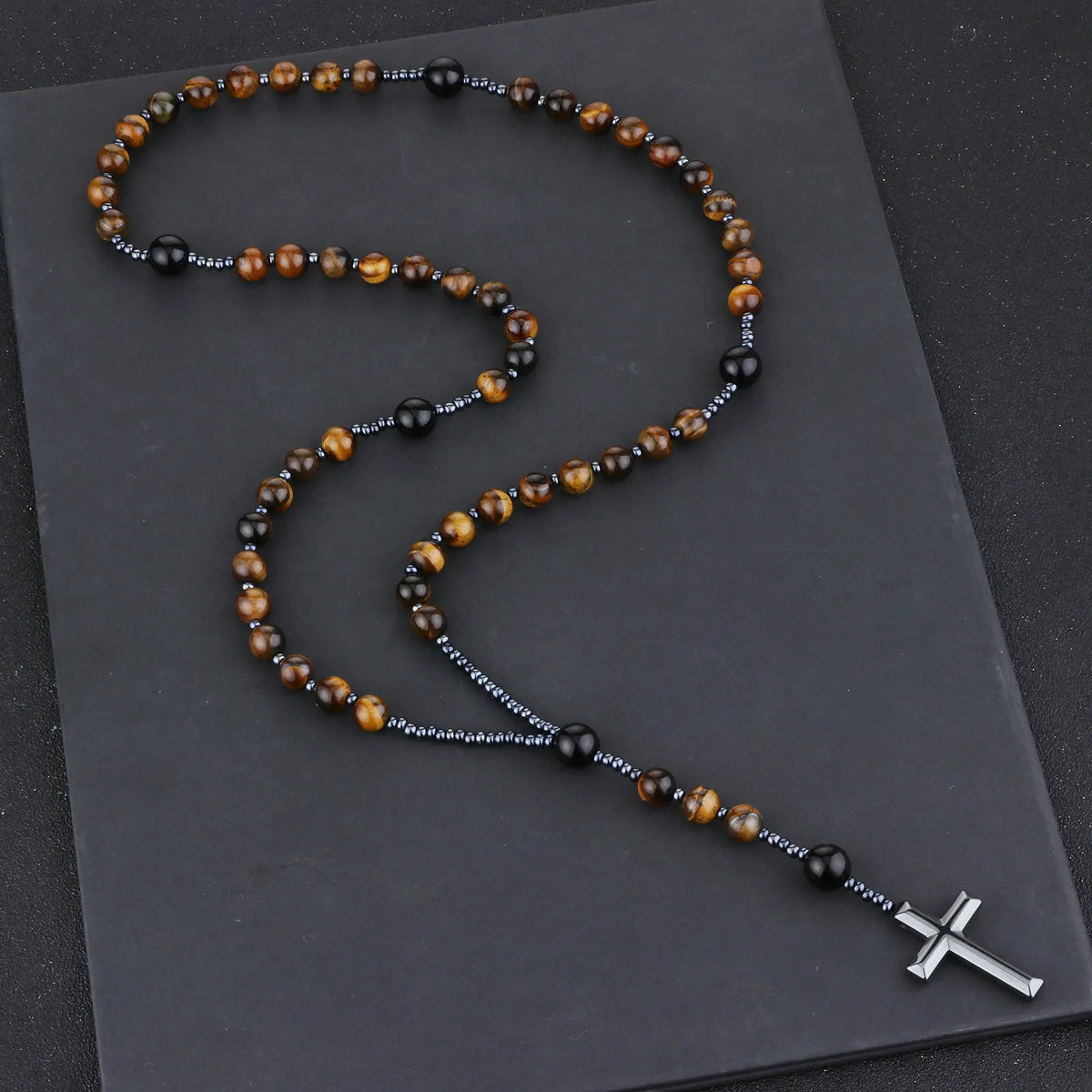 A rosary necklace featuring tiger eye and black agate beads with a hematite cross pendant on a dark gray background.