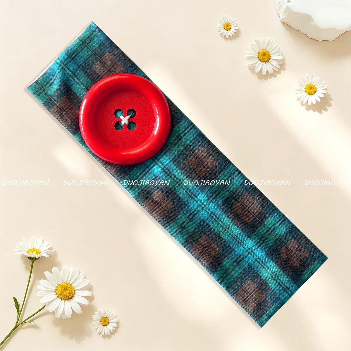 A teal plaid fabric headband with a red button attached, laid on a light background with white daisies.