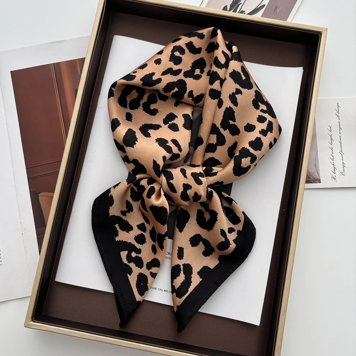 A black and tan leopard print silk scarf tied in a bow displayed in a wooden frame.