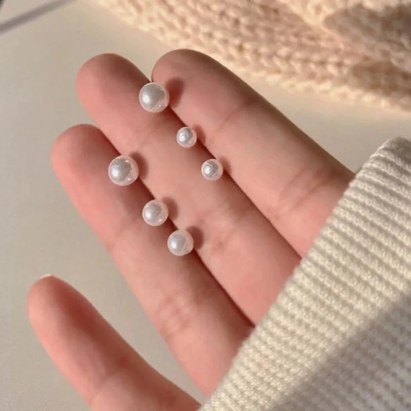 A hand holds three round, pearl-like stud earrings in gold-toned zinc alloy against a soft background.