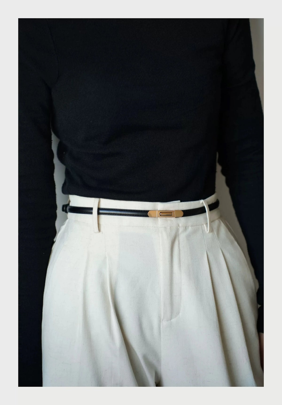 A close-up shot of a white pleated skirt and a black long-sleeved top paired with a thin gold buckle belt.