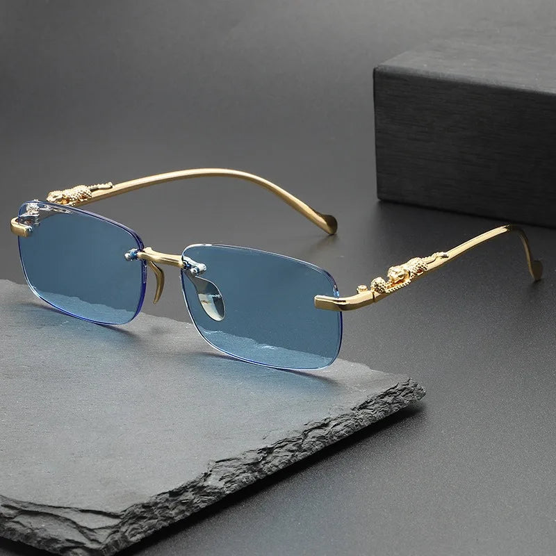 Rimless square sunglasses with gold-toned metal frames and blue mirrored lenses resting on a dark surface.