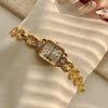 A gold-toned quartz fashion wristwatch with a square face and decorative links is shown on a light-colored fabric.