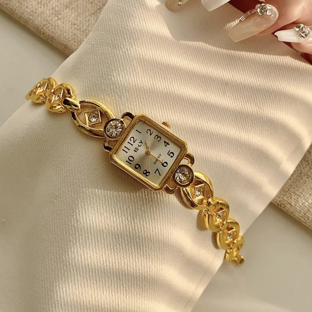 A gold-toned quartz fashion wristwatch with a square face and decorative links is shown on a light-colored fabric.
