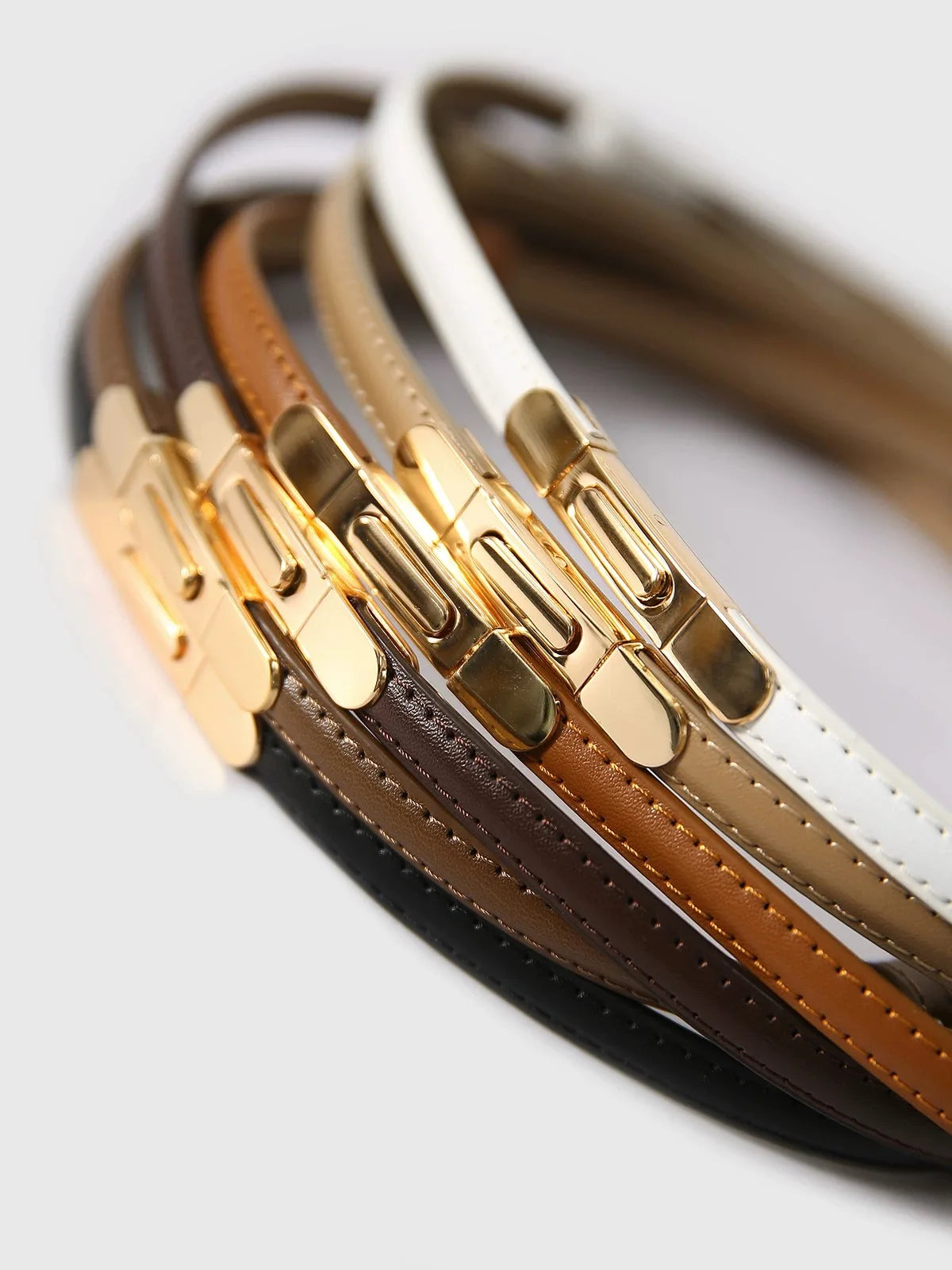 A close-up of several skinny leather belts in brown, tan, and white with gold buckles.