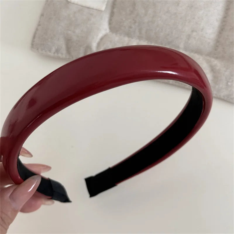 A close-up of a maroon PU leather fashion headband being held by a hand against a light background.