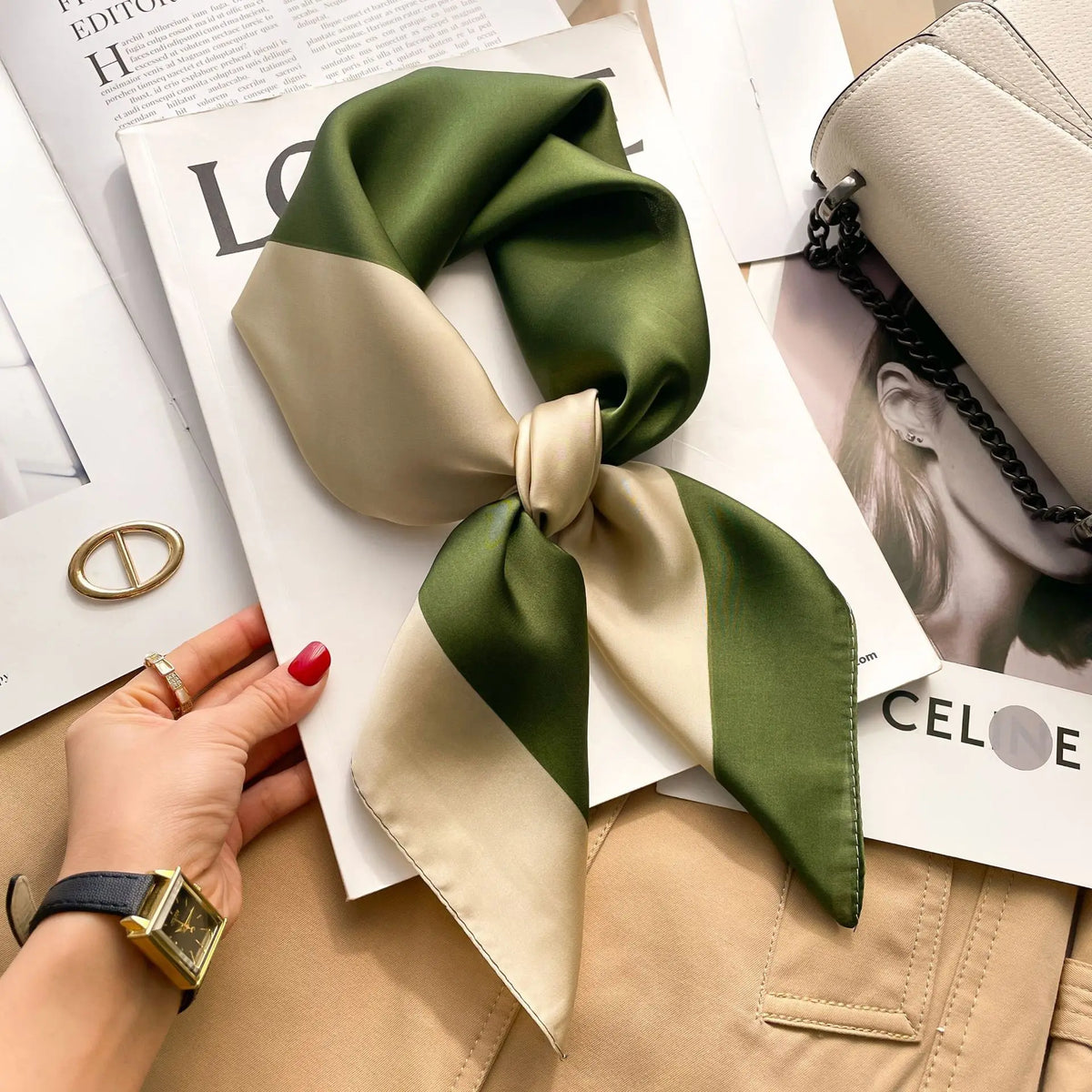 A luxury silk scarf in olive green and beige is tied into a bow, displayed on white paper next to a gold ring and watch.