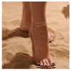 A close-up of a woman's ankle showing layered letter anklets on her skin against a sandy background.