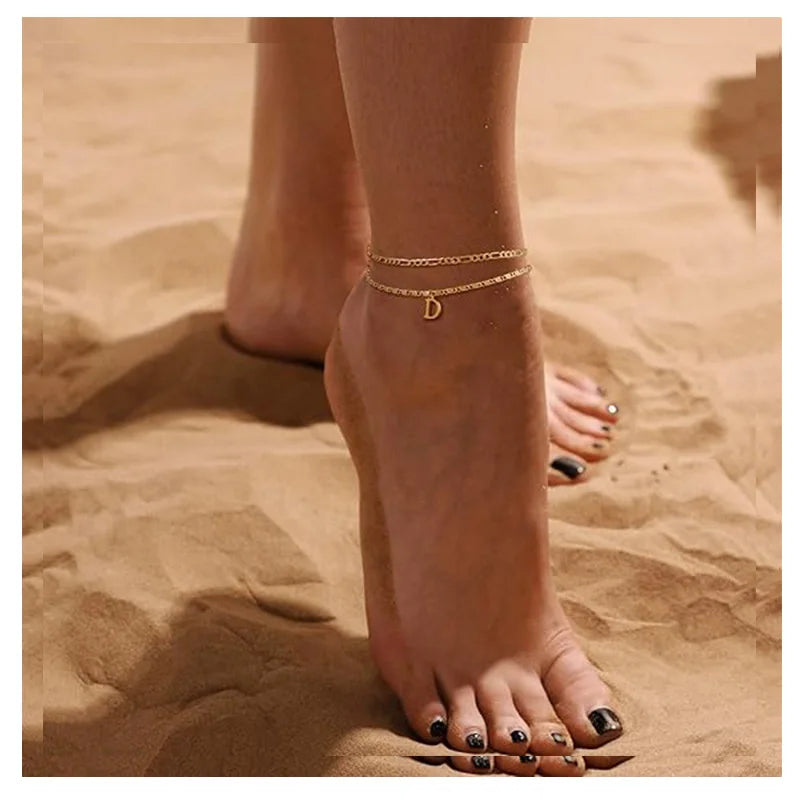 A close-up of a woman's ankle showing layered letter anklets on her skin against a sandy background.