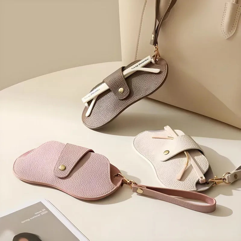A light pink PU leather eyewear storage case with a gold chain and white wooden sticks is displayed on a beige surface.