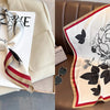 A cream silk scarf with a black floral print is displayed on a beige jacket and a brown leather sofa.