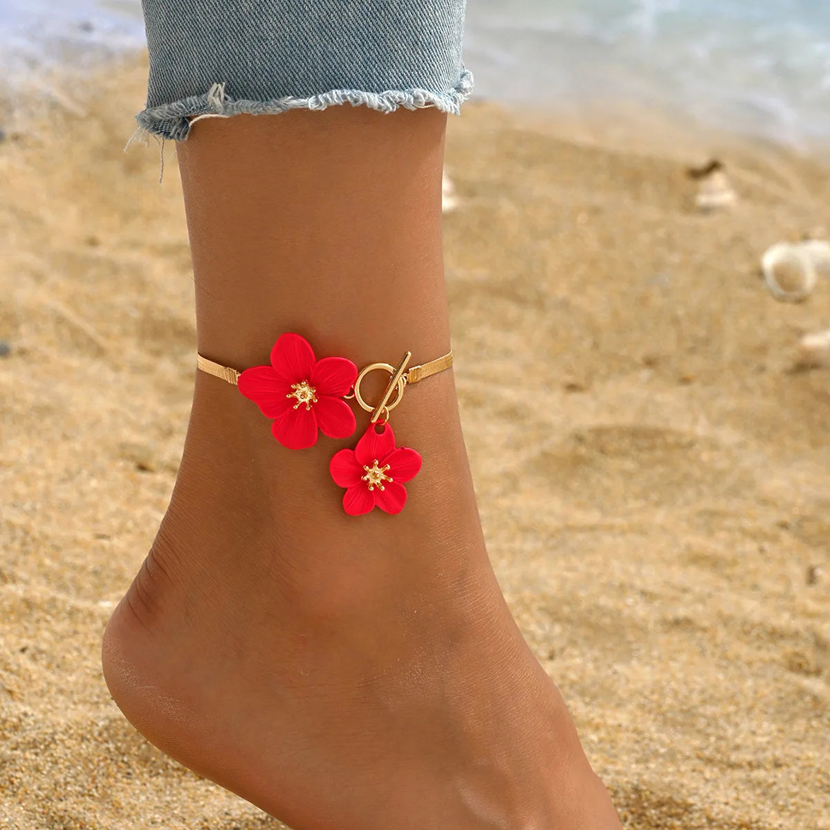 A close-up of a red floral bracelet worn on a person's ankle against a sandy beach background.