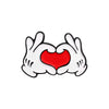 A white enamel pin depicting a cartoon hand gesture forming a heart shape in red.