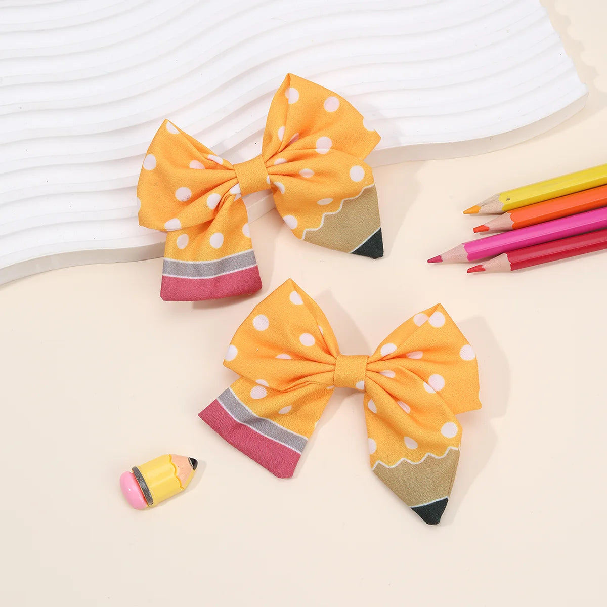 A set of cute pencil bow hair clips in orange and yellow with pink accents, displayed on a light background.