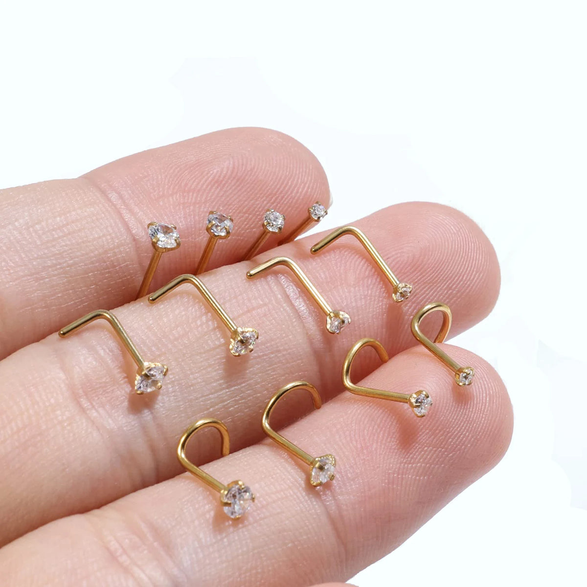 A hand holds several gold-finished stainless steel nostril studs with crystal accents.