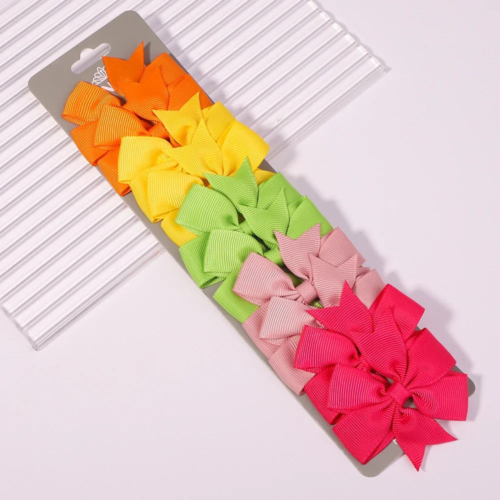 A collection of colorful handmade ribbon bow hair clips in orange, yellow, lime green, and pink arranged on a gray card.