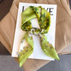 A light green silk scarf tied in a bow, displayed on a white card against a beige blazer.