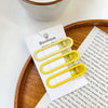 A set of vintage frosted geometry hair clips in yellow and gold are displayed on a wooden tray next to a piece of paper.