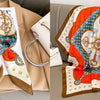 A floral print silk scarf featuring blue, orange, and white patterns displayed next to a light-colored leather bag.