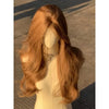 A long, wavy orange-brown wig displayed on a mannequin against a textured gray background.