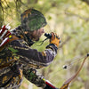 A person wearing a camouflage thermal beanie is aiming a bow and arrow outdoors.