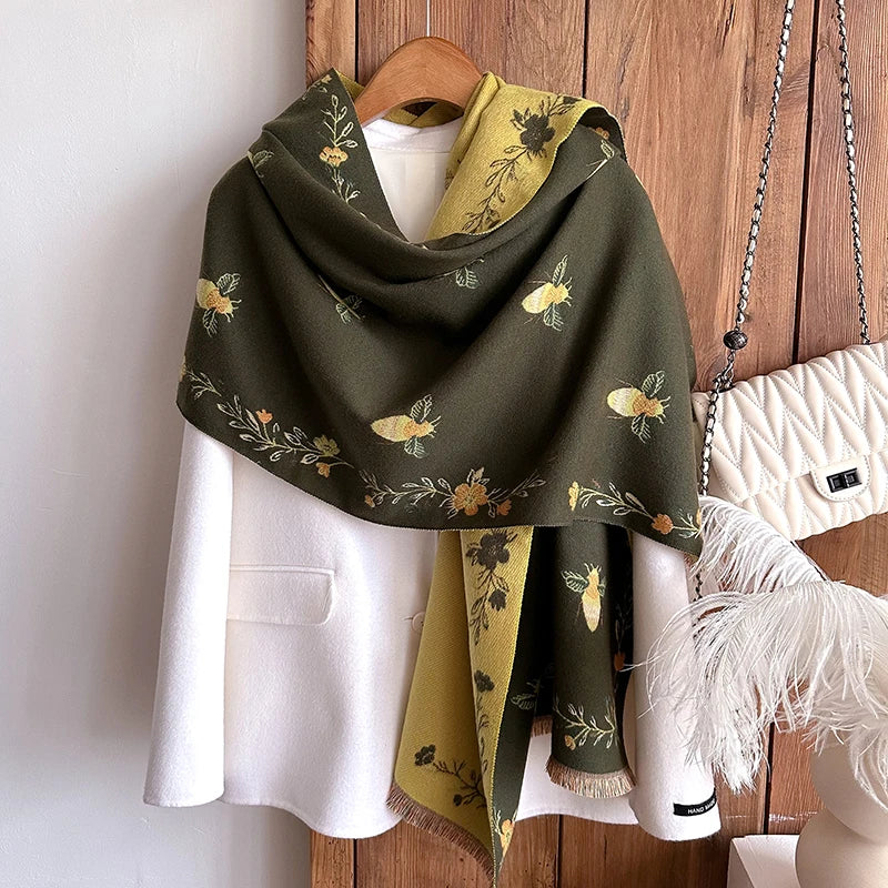 A dark olive green pashmina shawl with a floral print draped over a white blazer and displayed on a wooden hanger.