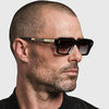 Mann trägt schwarze retro quadratische Metall-Sonnenbrille mit goldenen Akzenten vor hellem grauem Hintergrund.