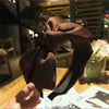 A dark brown chic wide bow headband is held up against a blurred indoor background.