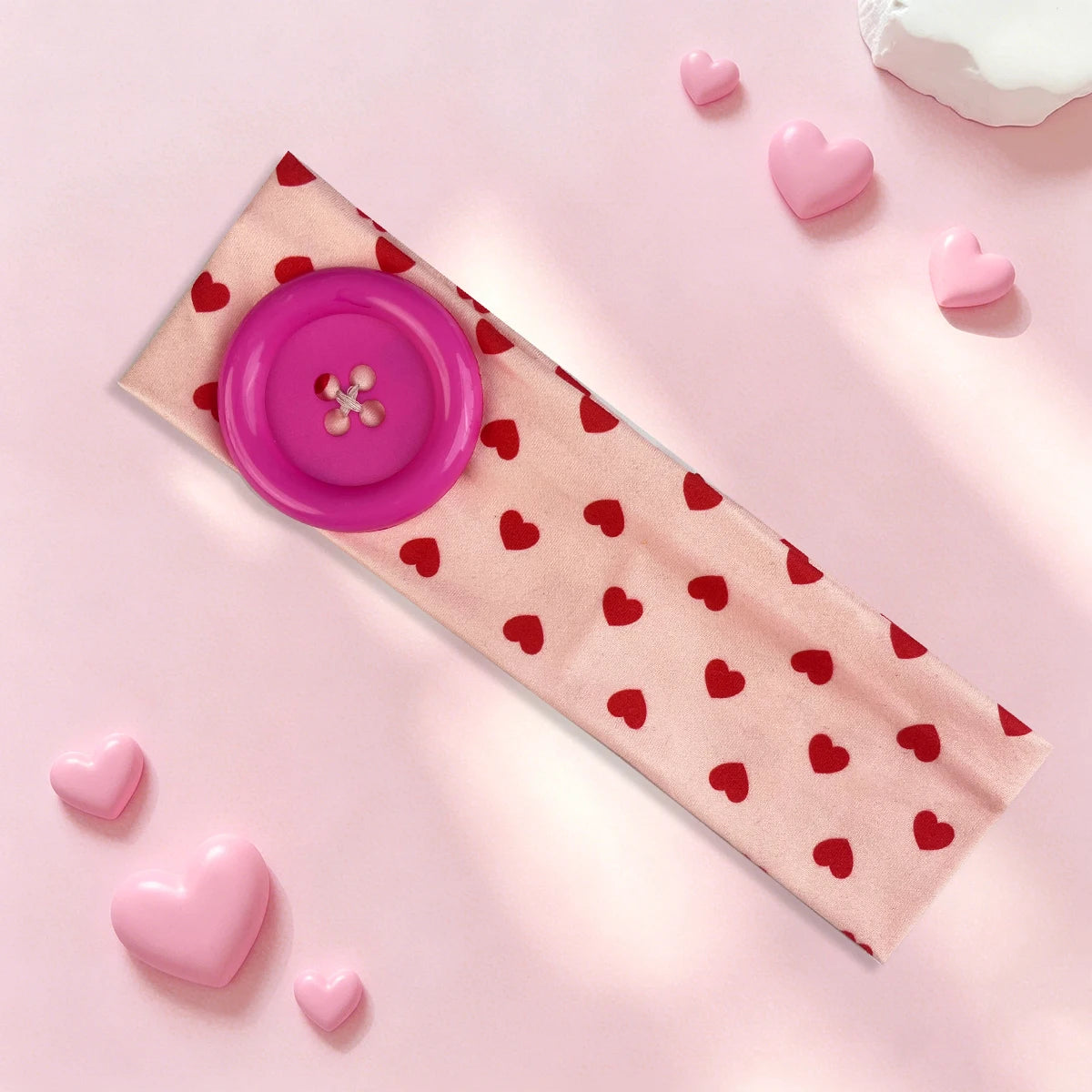 A pink vintage polka dot button headband lying on a light pink surface with small pink hearts scattered around it.