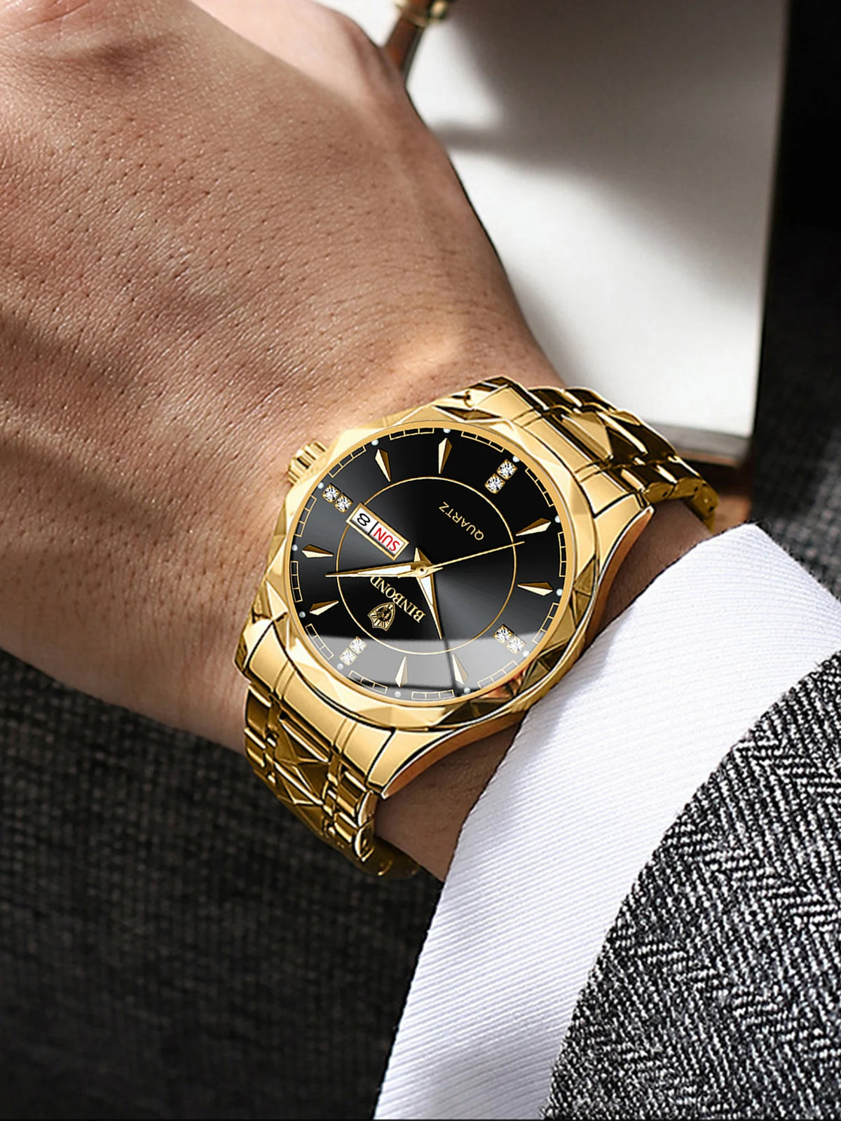 A close-up of a gold-toned stainless steel quartz watch worn on a wrist, paired with a white shirt and a textured suit sleeve