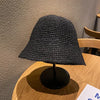 A black hand-woven straw visor hat displayed on a wooden surface against a gray wall.